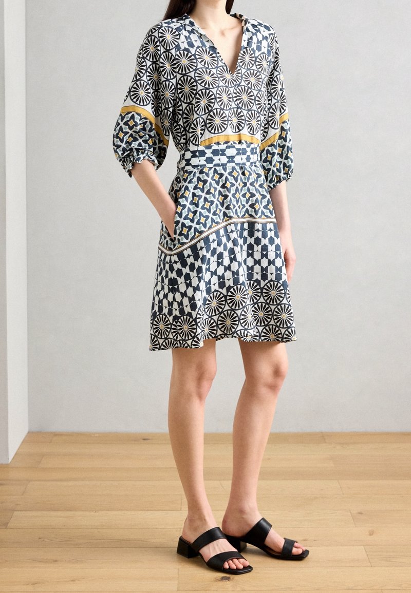 Femme portant une robe à motifs géométriques et floraux, avec des poches, des manches trois-quarts, et des sandales noires à talons sur un sol en bois.