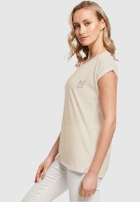 Beige kortærmet t-shirt lavet af bomuld, med et sommerfugl outline design på brystet. Glat tekstur og afslappet pasform.