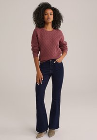 Pull en maille bordeaux avec texture en diamant, associé à un jean évasé bleu foncé et des bottes à talons taupe. Silhouette simple et moderne.