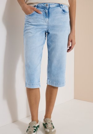 Pantalones cortos vaqueros azul claro hasta la rodilla, llevados por una persona con una mano en el bolsillo y zapatillas beige con detalles verdes.