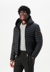 Svart dunjakke med hette, med glidelås og horisontal quiltet design. Båret over en grå skjorte og i kombinasjon med svarte bukser.