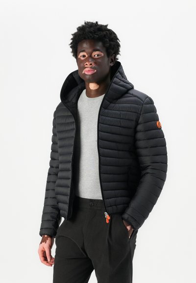 Doudoune noire avec capuche, dotée d'une fermeture éclair et d'un matelassage horizontal. Portée sur une chemise grise et associée à un pantalon noir.