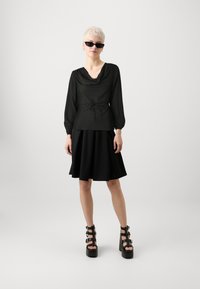 Blouse noire à col bénitier avec des manches longues et une ceinture à la taille, associée à une jupe noire évasée et des talons compensés noirs épais.