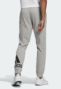 Person iført lysegrå Adidas sweatpants med sort logo nær anklen og hvide Adidas sneakers, stående mod en ensfarvet baggrund.