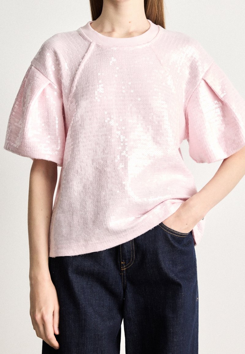 Femme portant un top à paillettes rose clair à manches courtes et un jean bleu foncé, une main dans la poche du jean, sur un fond uni.