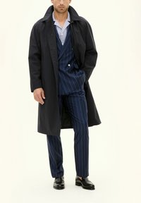 Langer schwarzer Mantel über einem blauen gestreiften Anzug mit einem gestreiften Hemd. Der Anzug hat eine taillierte Passform mit zwei Knöpfen. Schwarze Schuhe runden den Look ab.