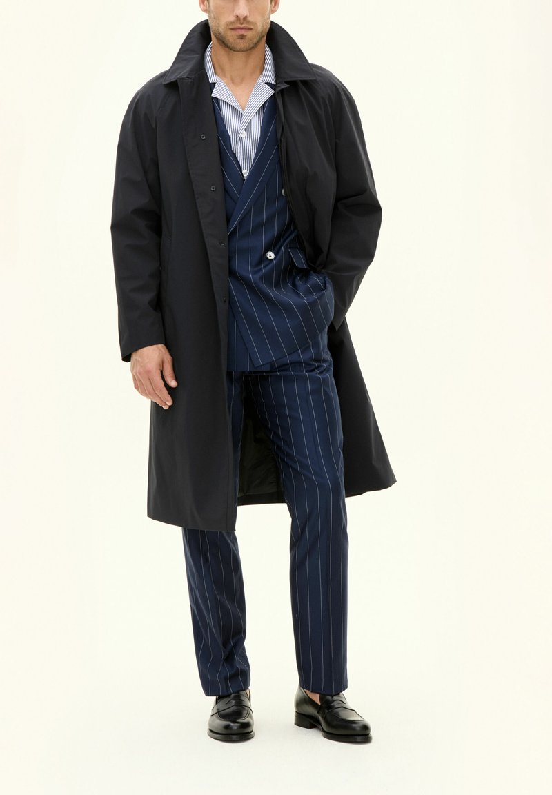 Langer schwarzer Mantel über einem blauen gestreiften Anzug mit einem gestreiften Hemd. Der Anzug hat eine taillierte Passform mit zwei Knöpfen. Schwarze Schuhe runden den Look ab.