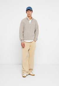 Nuori mies, joka on pukeutunut siniseen lippalakkiin, beigeen neulepaitaan valkoisen paidan päällä, leveisiin beigeihin housuihin ja valkoisiin tennareihin, seisoo valkoista taustaa vasten.