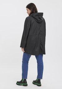 Black waterproof coat with a hood, featuring a relaxed fit and sleek texture. Paired with blue jeans and black-green ankle boots.
