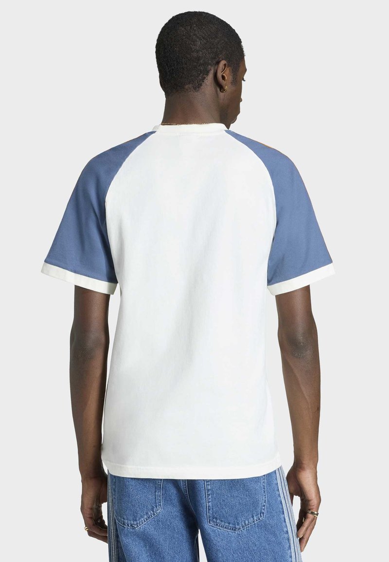 Mann trägt weißes T-Shirt mit blauen Raglanärmeln und hellblauen Jeans, von hinten vor einem einfarbigen Hintergrund gezeigt.