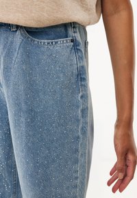 Jeans de mezclilla azul claro con tachuelas plateadas dispersas como embellecimiento. Diseño clásico de cinco bolsillos con un ligero ajuste holgado.