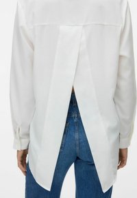 Hvid langærmet skjorte med en rygsplit-design, lavet af glat stof. Har knaplukninger ved ærmerne og en buet kant over denimjeans.