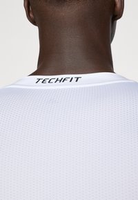 Vit sporttröja i ett texturerat, ventilerande tyg; har rundad hals och etikett med "TECHFIT" på baksidan, utformad för hög prestation.