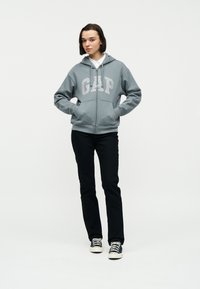 Femme debout les mains dans les poches, portant un sweat à capuche gris GAP, un pantalon noir, une chemise blanche à col, et des baskets noir et blanc, sur fond uni.