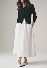 Sort kort cardigan, hvid flydende lang nederdel og lyse flade sko. Nederdelen har et tekstureret stof. Detaljerne inkluderer krydsede remme på skoene.