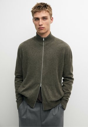 Junger Mann mit kurzen blonden Haaren trägt einen olivgrünen Pullover mit Reißverschluss und graue Hose, die Hände in den Taschen, vor einem schlichten Hintergrund.