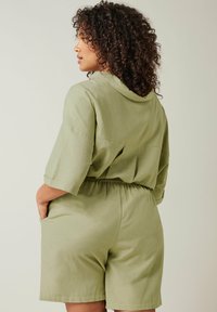 Khaki vihreä haalari, jossa on lyhyet hihat, kireä vyötärö ja sivutaskut. Kevyt kangas, jossa on sileä rakenne ja rento istuvuus.