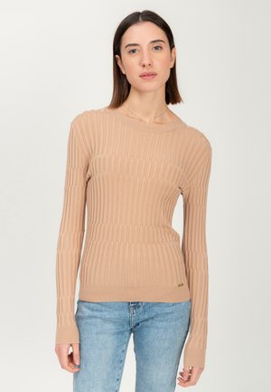 Vrouw in een beige geribbelde lange mouwen trui en lichtblauwe jeans, staand tegen een effen witte achtergrond.