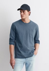 Langärmliges, blaues Baumwollhemd mit rundem Halsausschnitt und einer Brusttasche, kombiniert mit einer schwarzen Mütze, getragen mit hellblauen Jeans.