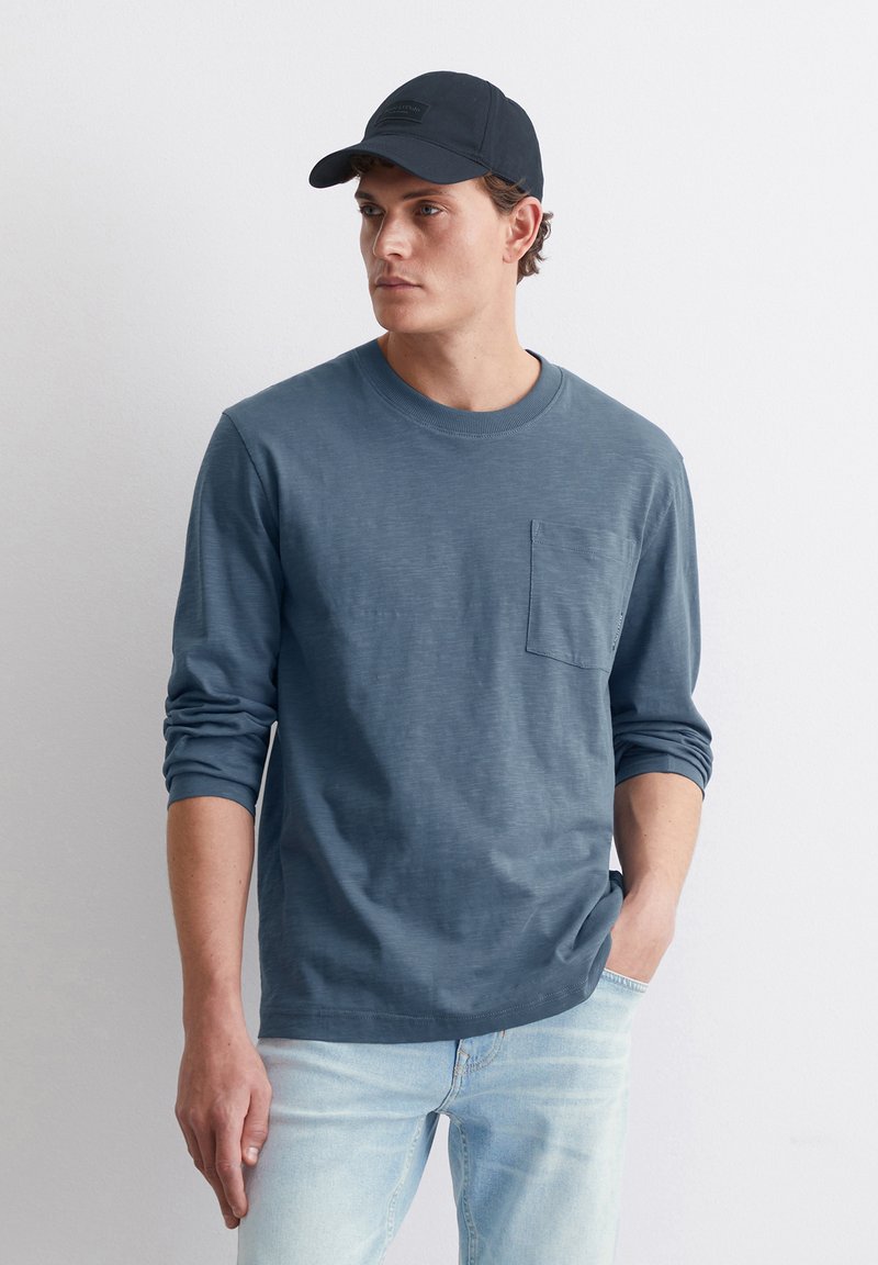 Langärmliges, blaues Baumwollhemd mit rundem Halsausschnitt und einer Brusttasche, kombiniert mit einer schwarzen Mütze, getragen mit hellblauen Jeans.