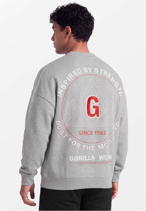 Man met krullend haar draagt een grijze trui met een rode "G" en fitness-geïnspireerde tekst op de rug, staand tegen een witte achtergrond.
