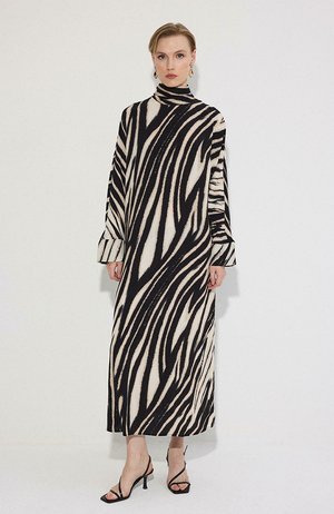 Vrouw die een zwart-wit zebraprint lange mouw coljurk en zwarte sandalen met hak en bandjes draagt, staand tegen een effen muur.