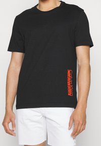 Svart bomull t-shirt med korta ärmar och rundad krage, med en vertikal orange logotryck på vänster sida.
