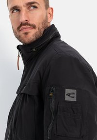 Black zip-up jacket with a high collar, featuring a zippered pocket on the left sleeve and textured accents. Durable fabric with a matte finish.