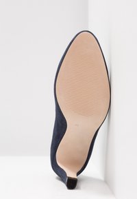 Chaussure à talon haut bleu marine vue de la semelle, reposant verticalement contre un mur d'angle blanc.