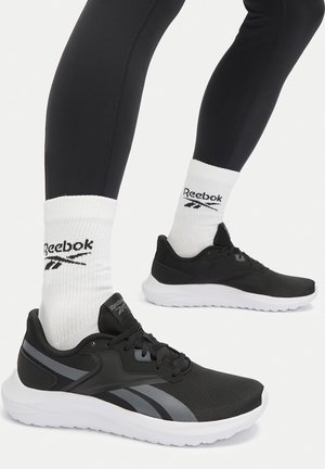 Piernas con mallas negras que llevan calcetines blancos Reebok hasta la tripa y zapatillas negras Reebok para correr con suelas blancas, de pie sobre un fondo blanco.