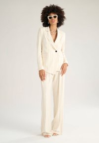 Completo a righe crema con giacca su misura e pantaloni a gamba larga, dotato di chiusura con un solo bottone e tessuto strutturato. Include occhiali da sole bianchi.