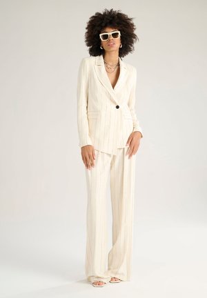 Costume crème rayé avec une veste ajustée et un pantalon large, présentant une fermeture à un bouton et un tissu texturé. Comprend des lunettes de soleil blanches.