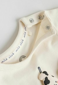 T-shirt en coton crème clair avec un motif de vache brodé. Comporte des boutons-pression au niveau de l'encolure et une texture de tissu douce et lisse.