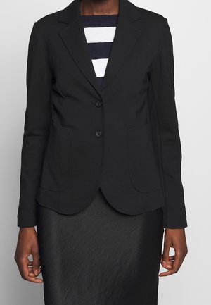 Persona que lleva una blazer negra ajustada con dos botones, bolsillos de parche y una camisa de rayas blancas y negras debajo, sobre un fondo liso.