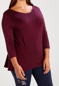 Burgunderfarbene Peplum-Bluse mit Dreiviertelärmeln, featuring einem runden Ausschnitt, verziert mit einem metallischen Akzent und floraler Stickerei an der Seite.