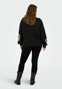 Zwarte gebreide trui met een losse fit en oversized mouwen, voorzien van een witte geborduurde afbeelding op elke arm, gecombineerd met zwarte skinny jeans en enkellaarsjes.
