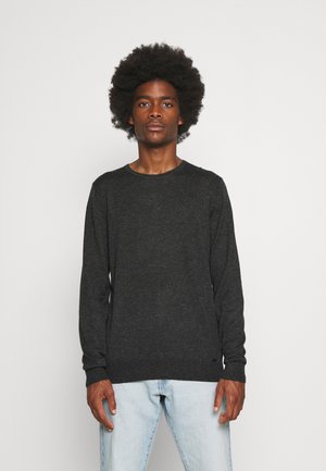 Junger Mann mit Afro-Frisur trägt dunkelgrauen langärmligen Pullover und hellblaue Jeans, steht vor einem schlichten weißen Hintergrund.