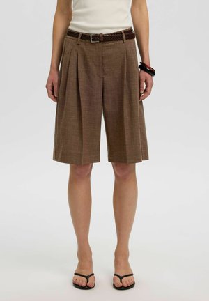 Femme portant un short plissé marron à hauteur du genou avec une ceinture tressée, un top blanc sans manches, des sandales noires et des bracelets foncés au poignet gauche.