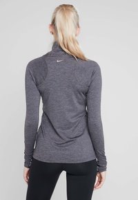 Haut de sport à manches longues pour femmes en tissu gris foncé chiné, avec col montant, passe-pouces et logo doré discret dans le dos.