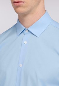 Ljusblå skjorta med knappar och spridd krage, med en slät textur och kontrasterande vita knappar.