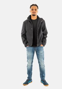 Giacca nera con zip, maglietta scura, jeans in denim azzurro chiaro e sneakers nere con dettagli blu. Il modello si trova in una posa neutra su uno sfondo bianco.