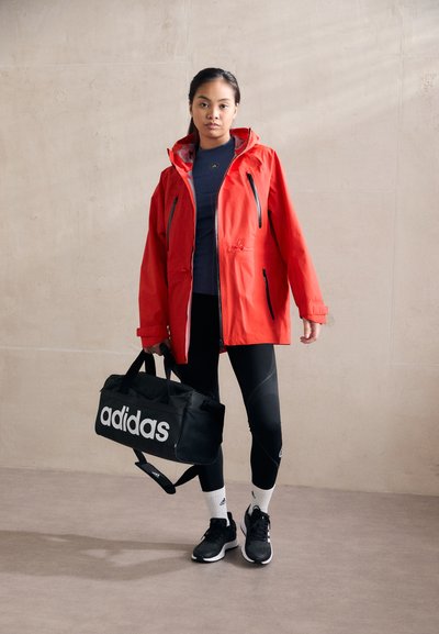 Red waterproof jacket with zippers, black leggings, and a black duffel bag with white "adidas" logo. Black sneakers with white soles.