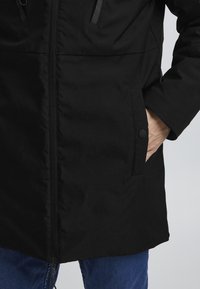 Parka neagră dintr-un material cu textură netedă, cu un fermoar central, buzunare laterale și detalii cu nasturi. Prim-plan pe jumătatea inferioară.