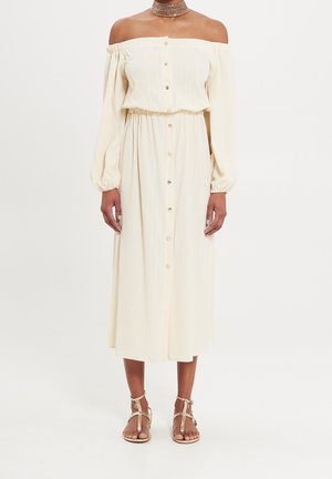 Woman wearing cream off-shoulder button-front midi dress with long sleeves, paired with gold strappy flat sandals and a multi-layered choker necklace.