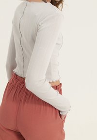 Mujer vestida con una camiseta de manga larga acanalada blanca y pantalones de tiro alto en color óxido con cinturilla elástica, con una mano en el bolsillo, de lado.