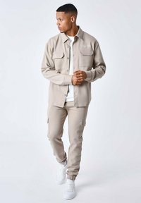 Beige fleece skjortejakke med to brystlommer, parret med matchende cargo-style bukser. Hvide sneakers fuldender outfittet.