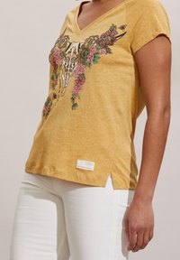 Mujer vestida con una camiseta amarilla mostaza de cuello en V con un diseño de flores y calaveras, combinada con pantalones blancos, de pie contra un fondo neutro.