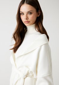 Biała dzianinowa sweter z ribbowanym kołnierzem szalowym, z wiązanym paskiem w talii i miękką strukturą. Design jest dopasowany w okolicy szyi i ramion.