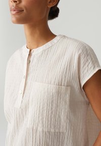 Camicia button-up di un delicato rosa, con texture, colletto alla mandarino, maniche corte e una singola tasca sul petto. Presenta sottili righe verticali.