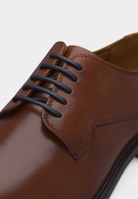 Καφέ γυαλιστερό δερμάτινο παπούτσι επίσημο με σκούρα μπλε κορδόνια και λεπτομερείς ραφές σε λευκό φόντο.